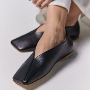 May include: Black leather flats with a square toe and a low heel. The shoes have a V-shaped vamp and a neutral-colored sole. The footwear is designed for comfort and style.