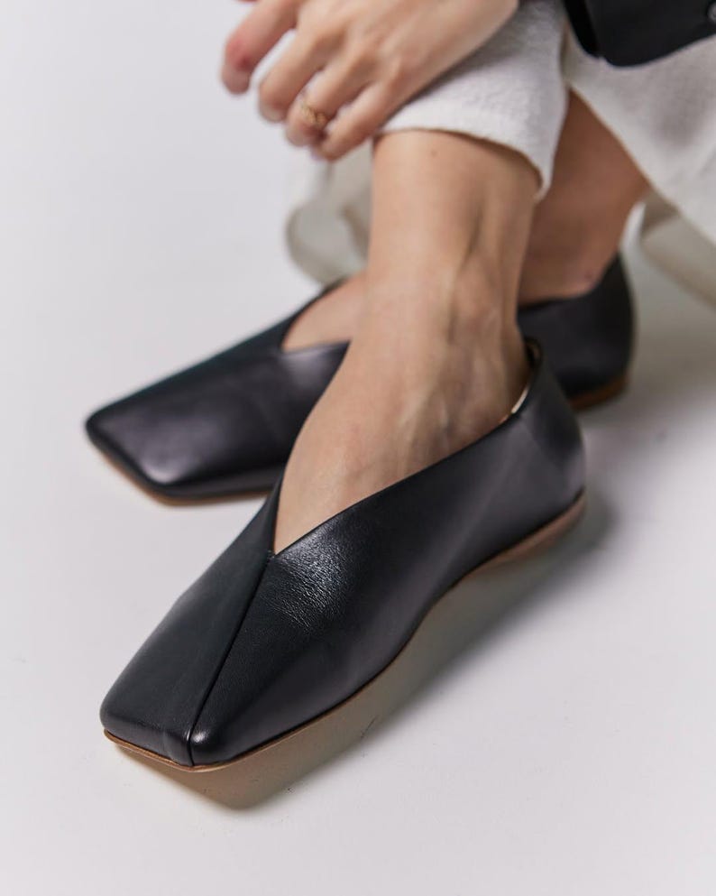 May include: Black leather flats with a square toe and a low heel. The shoes have a V-shaped vamp and a neutral-colored sole. The footwear is designed for comfort and style.