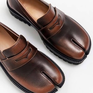 Pode incluir: Um par de mocassins de couro castanho com biqueira dividida. Os sapatos têm uma sola castanha escura e uma parte superior castanha mais clara com uma tira decorativa. O design da biqueira dividida é uma característica distintiva.