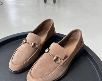 May include: A pair of brown suede loafers with a gold-colored metal buckle detail. The shoes have a rounded toe and a black sole. The loafers are displayed on a black surface.