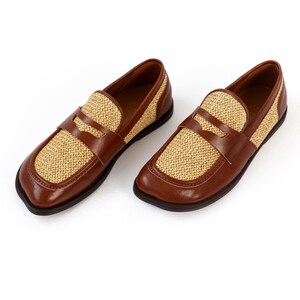 May include: A pair of brown leather loafers with woven tan accents. The shoes have a classic design with a rounded toe and a strap across the top. The soles are black.