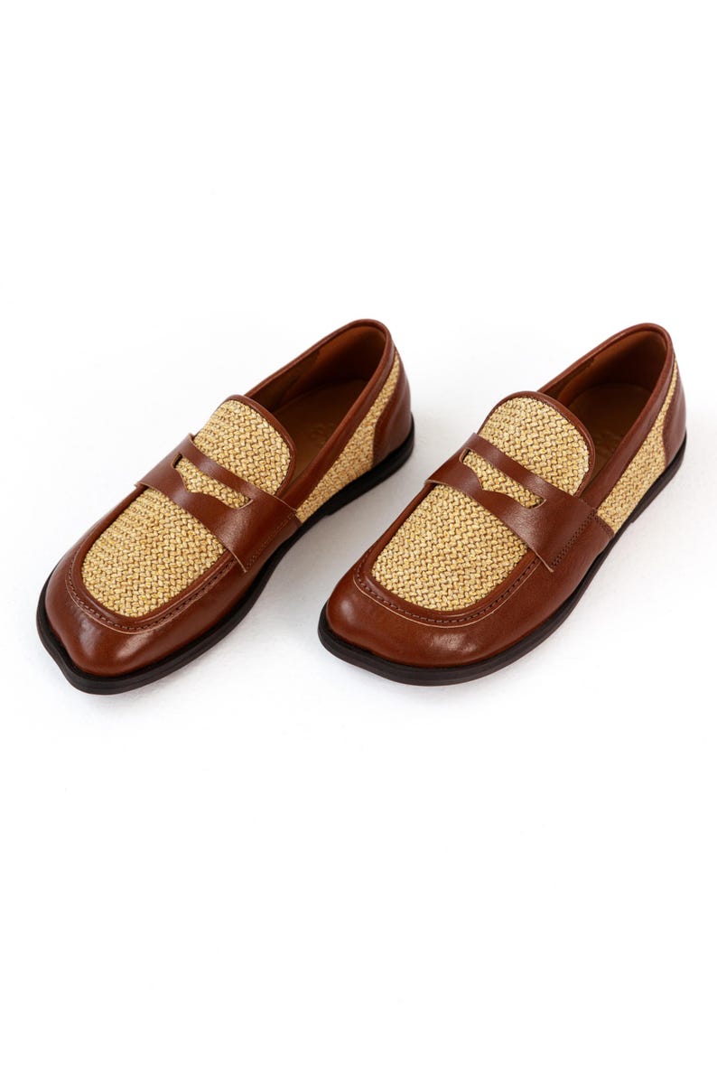 May include: A pair of brown leather loafers with woven tan accents. The shoes have a classic design with a rounded toe and a strap across the top. The soles are black.