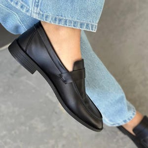 May include: Black leather loafers with a buckle detail. The loafers have a pointed toe and a low heel.