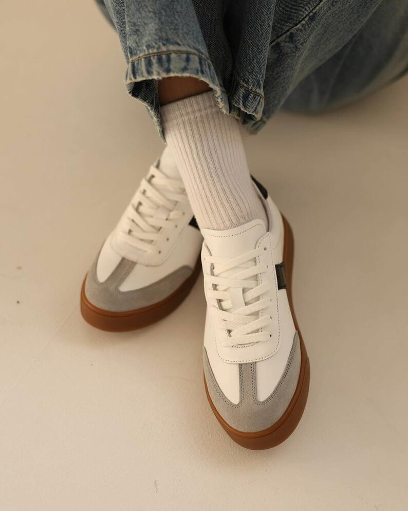 May include: White leather sneakers with a grey suede toe and a black stripe on the side. The sneakers have a gum sole.