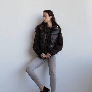 Op de afbeelding: Een persoon draagt een donkerbruin gewatteerd vest over een zwarte hoodie en lichtgrijze leggings. De outfit wordt gecompleteerd met donkerbruine enkellaarsjes met bontafwerking. De achtergrond is een effen witte muur.