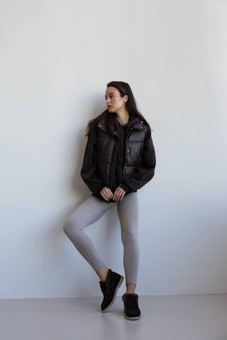 Op de afbeelding: Een persoon draagt een donkerbruin gewatteerd vest over een zwarte hoodie en lichtgrijze leggings. De outfit wordt gecompleteerd met donkerbruine enkellaarsjes met bontafwerking. De achtergrond is een effen witte muur.