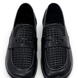 May include: A pair of black leather loafers. The shoes feature a woven design on the vamp and a strap across the top. The shoes are set against a white background.