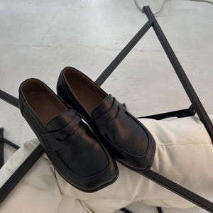 May include: A pair of black leather loafers with a square toe and a strap across the top. The shoes are displayed on a white surface, with a black metal frame in the background. The loafers have a classic, minimalist design.