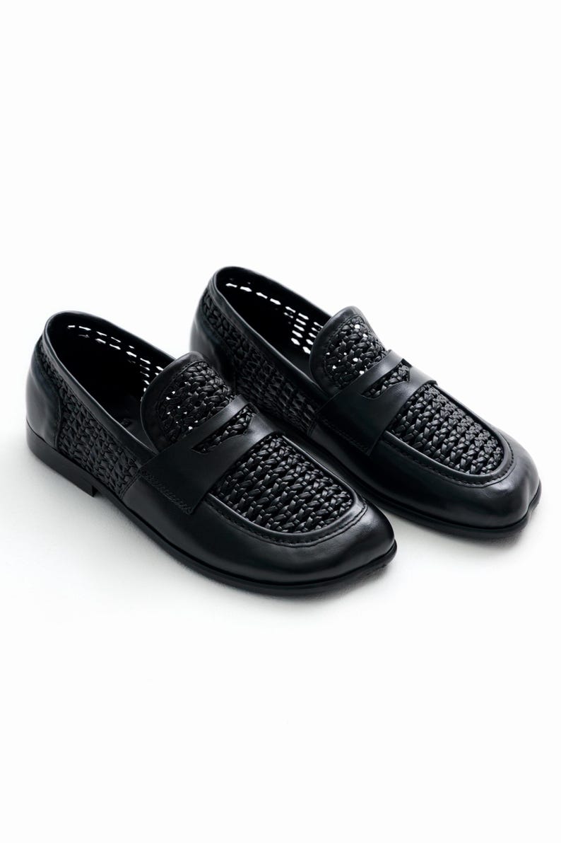 May include: A pair of black leather loafers with woven detailing on the vamp and sides. The shoes have a classic slip-on design with a low heel and a rounded toe. The shoes are set against a white background.