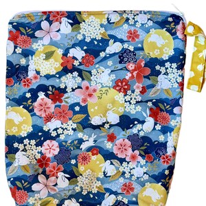 May include: A rectangular fabric wet bag featuring a dark blue background with a repeating pattern of white rabbits, yellow full moons, pink and red cherry blossoms, and small white floral clusters. It includes a white zip and a yellow handle.