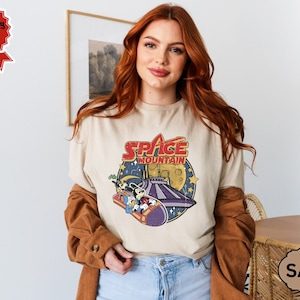 以下が含まれることがあります： ベージュのTシャツに、ミッキーマウスとその仲間たちがスペースマウンテンに乗っているヴィンテージスタイルのグラフィックがプリントされています。 「スペースマウンテン」の文字は赤と青で印刷されています。