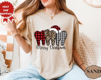 Op de afbeelding: Beige T-shirt met drie decoratieve tanden met feestelijke patronen: rood-zwarte buffelruit, luipaardprint en zwart-wit geruit. Een kerstmuts siert een tand. De woorden "Merry Christmas" staan eronder.