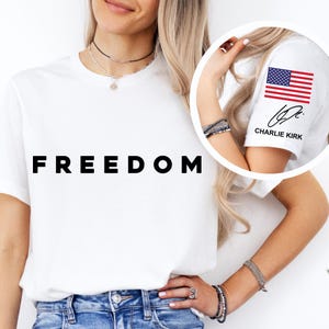 May include: White t-shirt with the word "FREEDOM" in large, black capital letters. The sleeve features an American flag graphic and the name "CHARLIE KIRK". The shirt is worn by a person.