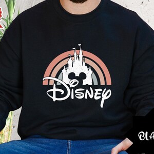 Puede incluir: Una sudadera negra de manga larga con un gr&aacute;fico blanco de la silueta de un castillo dentro de un arco&iacute;ris de tonos apagados. Debajo del castillo, la palabra Disney est&aacute; impresa en una fuente cursiva blanca. El dise&ntilde;o tiene una textura desgastada.