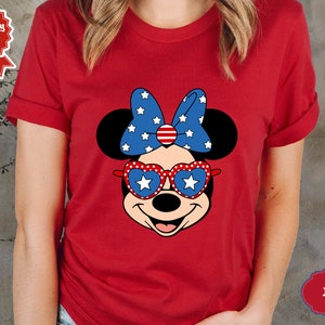 May include: Red t-shirt with a cartoon image of Minnie Mouse wearing red, white, and blue heart-shaped sunglasses and a bow with stars and stripes.