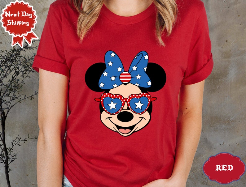 May include: Red t-shirt with a cartoon image of Minnie Mouse wearing red, white, and blue heart-shaped sunglasses and a bow with stars and stripes.