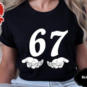 May include: Black t-shirt with the number 67 in large white font. Below the numbers are two open hands in a white line drawing. The shirt has a round neck and short sleeves. The words "Next Day Shipping" are in a red and white emblem.
