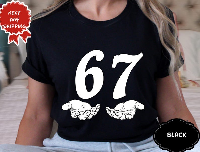 May include: Black t-shirt with the number 67 in large white font. Below the numbers are two open hands in a white line drawing. The shirt has a round neck and short sleeves. The words "Next Day Shipping" are in a red and white emblem.