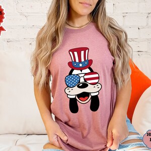 May include: A pink t-shirt with a cartoon dog wearing a red, white, and blue hat and sunglasses. The sunglasses have the American flag design.