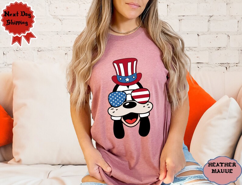 May include: A pink t-shirt with a cartoon dog wearing a red, white, and blue hat and sunglasses. The sunglasses have the American flag design.