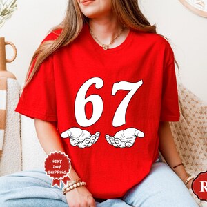May include: A red t-shirt with the number 67 in white, above a pair of open hands. The shirt is a solid red color. The text "RED" is in a red oval on the bottom right of the shirt.