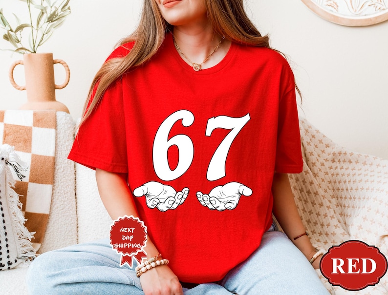 May include: A red t-shirt with the number 67 in white, above a pair of open hands. The shirt is a solid red color. The text "RED" is in a red oval on the bottom right of the shirt.