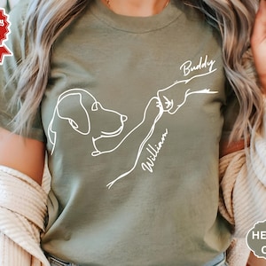 Op de afbeelding: Heather olijfgroen t-shirt met een witte lijntekening van een hond die een hand aanraakt. De namen "Buddy" en "William" staan boven en onder de hand geschreven. Het shirt heeft een ronde hals en korte mouwen.