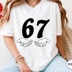 May include: White t-shirt featuring the number 67 in bold black font, with two stylized hands beneath. The shirt is a simple design, perfect for casual wear. The image also includes a "Next Day Shipping" graphic.