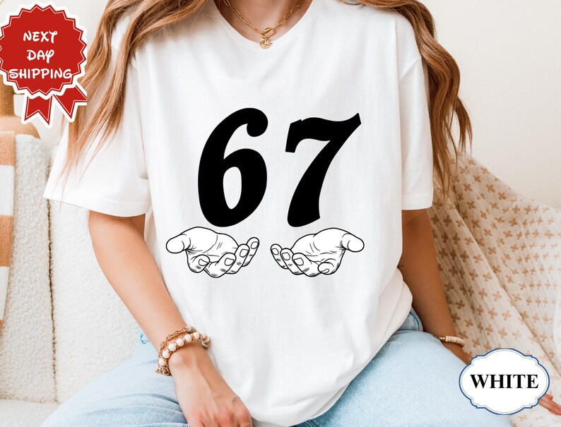 May include: White t-shirt featuring the number 67 in bold black font, with two stylized hands beneath. The shirt is a simple design, perfect for casual wear. The image also includes a "Next Day Shipping" graphic.