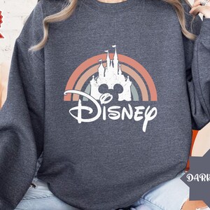 Puede incluir: Una sudadera de manga larga gris oscuro con un gr&aacute;fico blanco de la silueta de un castillo sobre un arco&iacute;ris de colores apagados. La palabra Disney est&aacute; impresa en una fuente cursiva blanca debajo del dise&ntilde;o del castillo y el arco&iacute;ris.