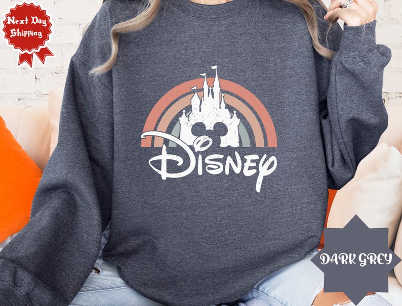 Puede incluir: Una sudadera de manga larga gris oscuro con un gr&aacute;fico blanco de la silueta de un castillo sobre un arco&iacute;ris de colores apagados. La palabra Disney est&aacute; impresa en una fuente cursiva blanca debajo del dise&ntilde;o del castillo y el arco&iacute;ris.