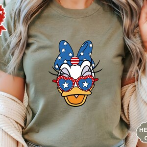 May include: A green t-shirt with a cartoon image of a duck wearing red, white, and blue sunglasses and a bow. The duck has a white star on its sunglasses and a red, white, and blue bow with white stars.