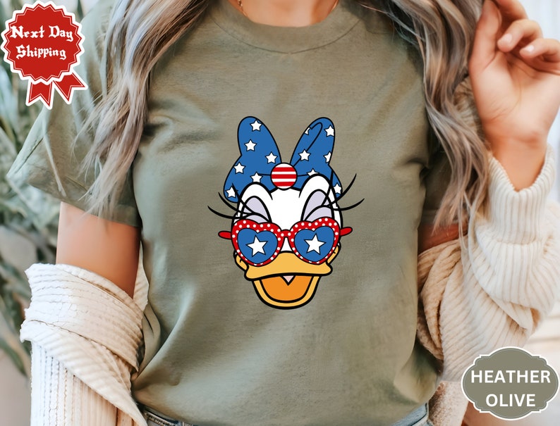 May include: A green t-shirt with a cartoon image of a duck wearing red, white, and blue sunglasses and a bow. The duck has a white star on its sunglasses and a red, white, and blue bow with white stars.