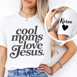 May include: White t-shirt with the text "cool moms love Jesus" in a dark gray, distressed font. The sleeve of the shirt has the name "Karen" and a black heart. The shirt is worn by a person.