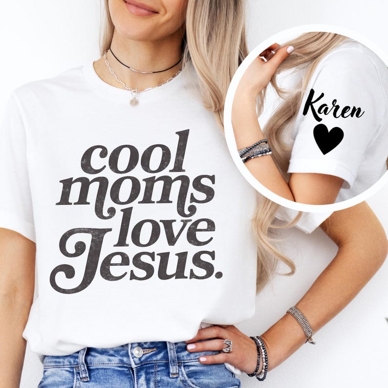 May include: White t-shirt with the text "cool moms love Jesus" in a dark gray, distressed font. The sleeve of the shirt has the name "Karen" and a black heart. The shirt is worn by a person.