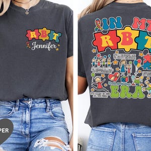 以下が含まれることがあります： カラフルなパズルピースのデザインが施された濃いグレーのTシャツ。「RBT」と「Jennifer」の文字が前面にプリントされています。背面には「IN MY ERA」の文字とパズルピース、イラストが描かれています。普段使いにぴったりのカジュアルなTシャツです。