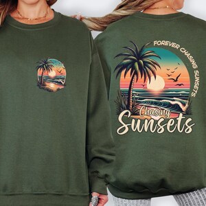 May include: Dark green sweatshirt featuring a sunset scene with palm trees, waves, and birds. The back of the sweatshirt has the text "Forever Chasing Sunsets" and "Chasing Sunsets" below the graphic.