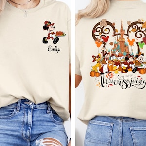 May include: A beige t-shirt with a Mickey Mouse character dressed as a pilgrim holding a plate of food. The text "Emily" is printed below the character. The back of the shirt features a Mickey Mouse head silhouette with a Thanksgiving scene inside. The scene includes Mickey Mouse and other Disney characters dressed in Thanksgiving attire, a castle, and the text "Happy Thanksgiving".