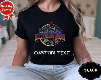 May include: Black t-shirt featuring a colourful graphic of the Universal Studios logo and various characters, including a dinosaur, a minion, and a Hogwarts castle. The text "CUSTOM TEXT" is printed below the graphic.