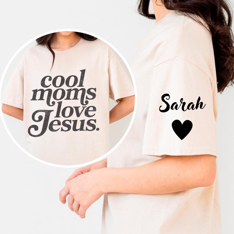 May include: A beige t-shirt with the text "cool moms love Jesus" in black script on the front. The right sleeve has the name "Sarah" in script with a black heart below it. The shirt is a casual, everyday wear item.