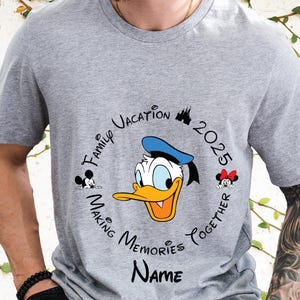 May include: Grey t-shirt with a circular design featuring Donald Duck in a blue sailor hat. The design includes the text "Family Vacation 2025 Making Memories Together" and a space for a name.