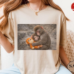 May include: A beige t-shirt featuring a printed image of two monkeys embracing, one holding an orange stuffed monkey. The image is set against a gray background. The shirt has short sleeves and a round neckline.