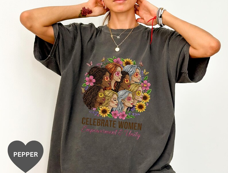 May include: Dark gray t-shirt featuring a floral design with multiple women's faces, sunflowers, and butterflies. The text "CELEBRATE WOMEN Empowerment & Unity" is printed below the design. The shirt has short sleeves.