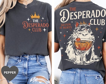 Può includere: Una t-shirt grigio antracite con una grafica frontale raffigurante una corona e il testo The Desperado Club. La grafica sul retro mostra un gatto con occhiali da sole e una corona, che tiene un cocktail con una ciliegia, e il testo The Desperado Club So Fun It Hurts.