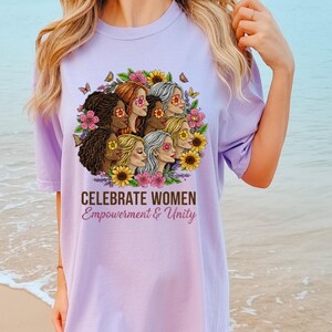 May include: A light purple t-shirt featuring a graphic of diverse women surrounded by flowers and butterflies. The text "CELEBRATE WOMEN Empowerment & Unity" is printed below the graphic. The shirt is a comfortable, casual style.