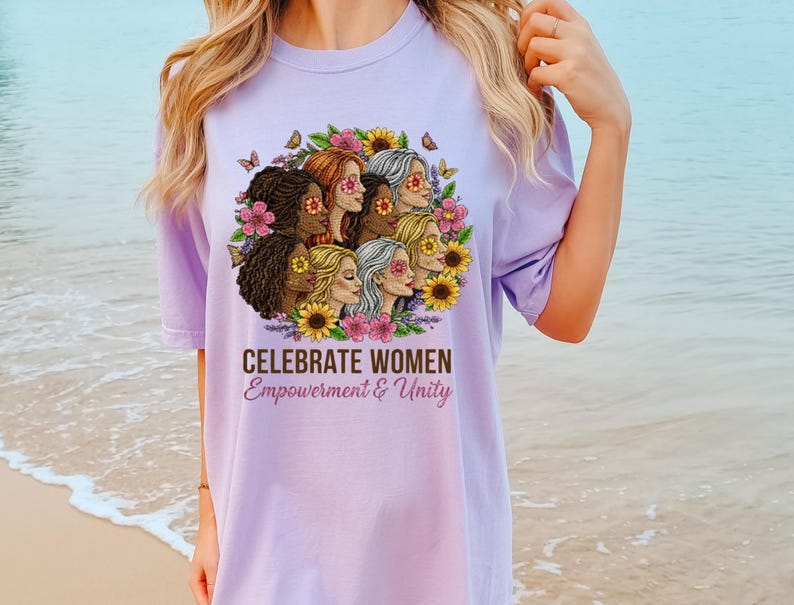 May include: A light purple t-shirt featuring a graphic of diverse women surrounded by flowers and butterflies. The text "CELEBRATE WOMEN Empowerment & Unity" is printed below the graphic. The shirt is a comfortable, casual style.