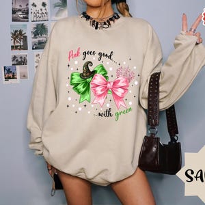 May include: Sand-colored sweatshirt with a graphic featuring a green and pink bow design, a witch hat, and the text "Pink goes good...with green." The sweatshirt has long sleeves and a crew neck.