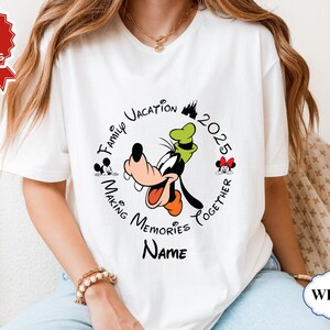 May include: White t-shirt with a circular design. The design includes the text "Family Vacation 2025 Making Memories Together" and cartoon images of Goofy, Mickey, and Minnie Mouse. The word "Name" is below the design.