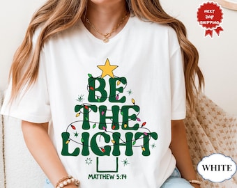 Peut inclure: T-shirt blanc avec un motif de sapin de Noël. Le motif présente les mots "BE THE LIGHT" en vert, avec une étoile jaune en haut et des lumières colorées. Le texte "MATTHIEU 5:14" est en bas.