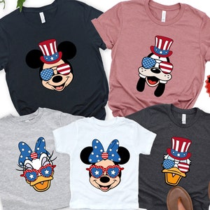 May include: Set of five t-shirts featuring cartoon characters wearing patriotic themed hats and sunglasses. The characters include Mickey Mouse, Minnie Mouse, Donald Duck, and Goofy. The shirts are in various colors, including navy blue, gray, white, and pink.